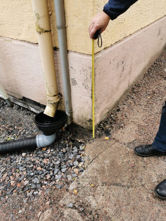Kuvan tarkoitus on dokumentoida pihan korko- ja kuivatusongelma, jossa sadevesi pääsee kulkeutumaan sokkelia vasten. Mittaus osoittaa korjaustarpeen ja perustelee hulevesiratkaisujen suunnittelun työmaalla.
